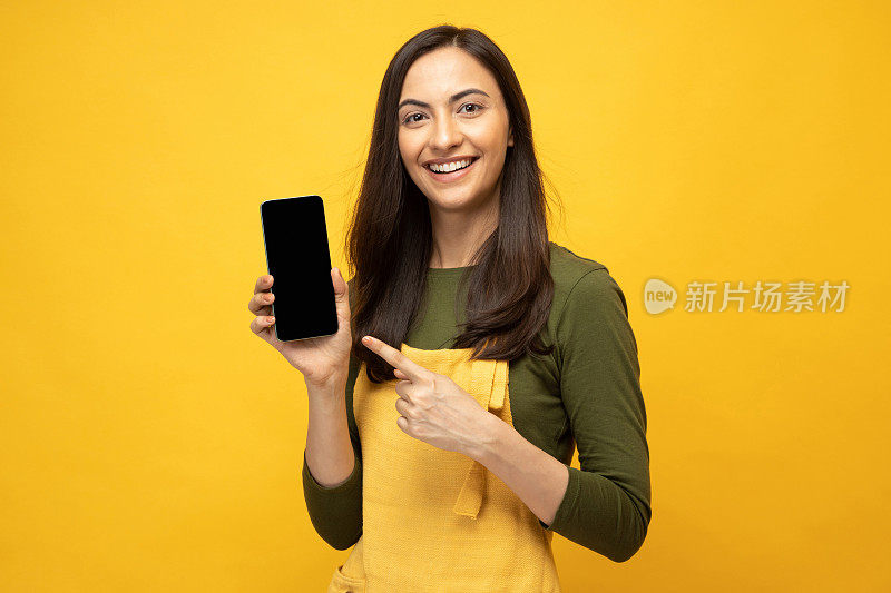Photo of pretty positive young woman showing device screen in yellow apron isolated on yellow background stock photo